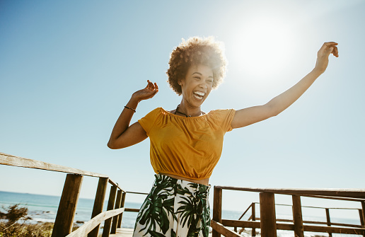 Excited young woman running on a boardwalk with her hands raised on a sunny day. African female having fun on summer vacation at the seaside.