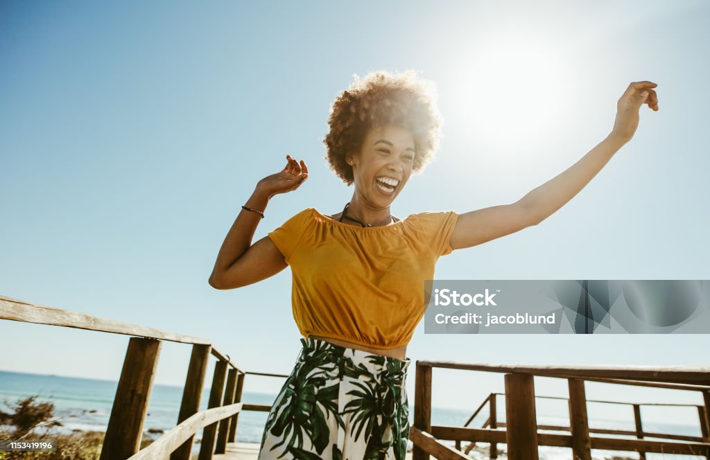 Woman having fun on summer vacation Excited young woman running on a boardwalk with her hands raised on a sunny day. African female having fun on summer vacation at the seaside. Women Stock Photo