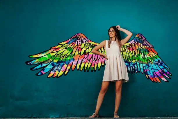 Smiling Caucasian girl in white summer dress posing in front of turquoise wall with colorful wings.