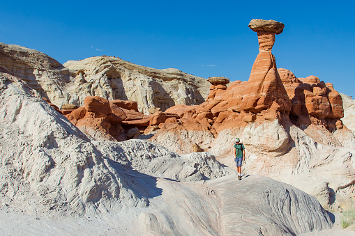 A boy walks along the top of white sandstone with a tall red hoodoo behind him.