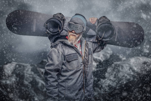 istock Brutal redhead snowboarder with a full beard in a winter hat and protective glasses dressed in a snowboarding coat holds a snowboard on his shoulder in a studio. 1153429115