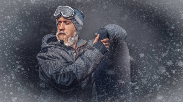 istock Brutal redhead snowboarder with a full beard in a winter hat and protective glasses dressed in a snowboarding coat posing with snowboard at a studio, looking away. 1153429255