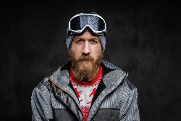 istock Confident man with a red beard wearing a full equipment for extreme snowboarding, looking at a camera with a serious look, isolated on a dark textured background. 1153429308
