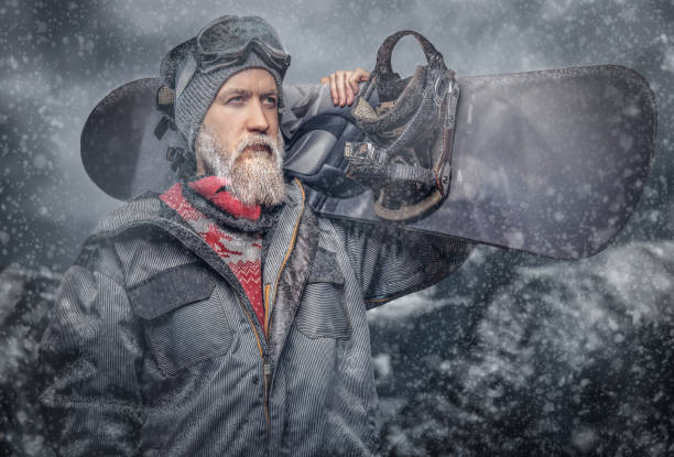 istock Brutal redhead snowboarder with a full beard in a winter hat and protective glasses dressed in a snowboarding coat posing with snowboard at a studio, looking away. 1153429349