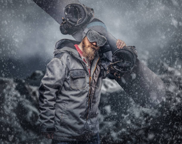 istock Brutal redhead snowboarder with a full beard in a winter hat and protective glasses dressed in a snowboarding coat holds a snowboard on his shoulder in a studio 1153429371
