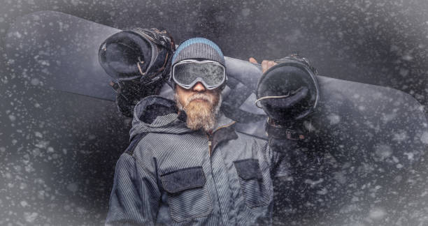 istock Brutal redhead snowboarder with a full beard in a winter hat and protective glasses dressed in a snowboarding coat posing with snowboard at a studio. 1153429391
