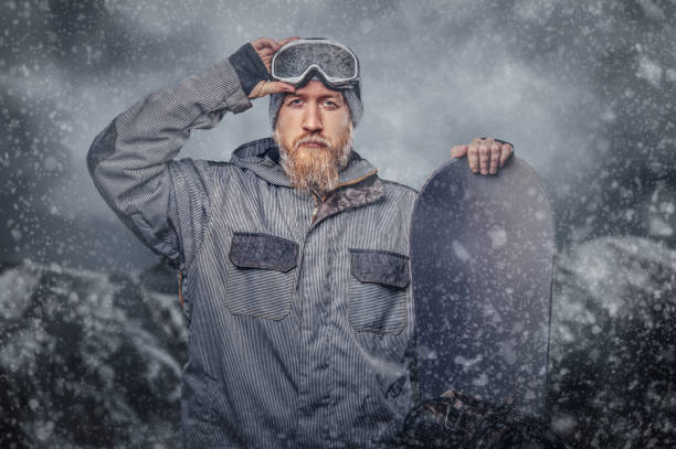 istock Brutal redhead snowboarder with a full beard in a winter hat and protective glasses dressed in a snowboarding coat posing with snowboard at a studio. 1153430504