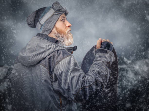 istock Brutal redhead snowboarder with a full beard in a winter hat and protective glasses dressed in a snowboarding coat posing with snowboard at a studio, looking away. 1153430530