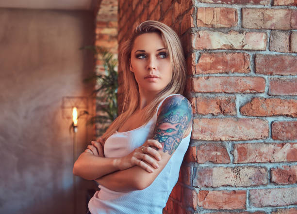 porträt einer blonden hipster-frau, die mit gekreuzten armen steht, während sie sich an eine ziegelwand in einem raum mit einem loft-innenraum lehnt. - tattoo t shirt brick wall portrait stock-fotos und bilder