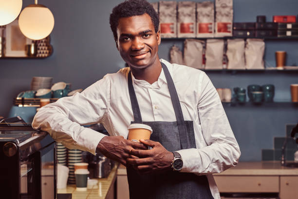 istock African barista smiling at camera relaxing after workday with coffee while leaning on the counter at coffee shop. 1153440248