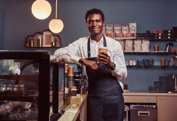 istock African barista smiling at camera relaxing after workday with coffee while leaning on the counter at coffee shop. 1153440296