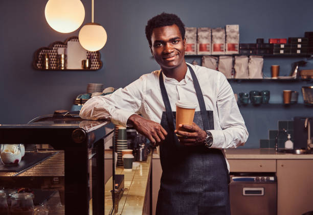 istock African barista smiling at camera relaxing after workday with coffee while leaning on the counter at coffee shop. 1153440344