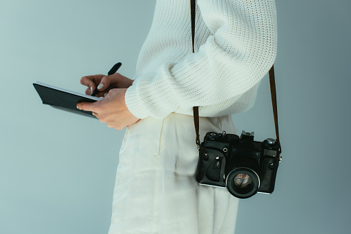 cropped view of young woman writing in notebook while standing with digital camera on grey
