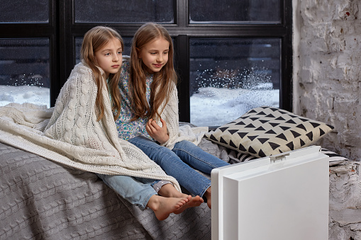 Two beautiful caucasian little sisters sitting on the windowsill, covered by blanket. They froze up and trying to warm up, using heater near their foots