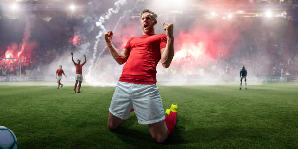 Professional Soccer Player Celebrating On Knees In Stadium With Flares Close up image of a professional male soccer player kneeling on a pitch with fists clenched, arms raised, screaming with open mouth in celebration of victory. The footballer is wearing a plain red top and white shorts. Flares are being let off by spectators in the stands in the background. soccer player photos stock pictures, royalty-free photos & images