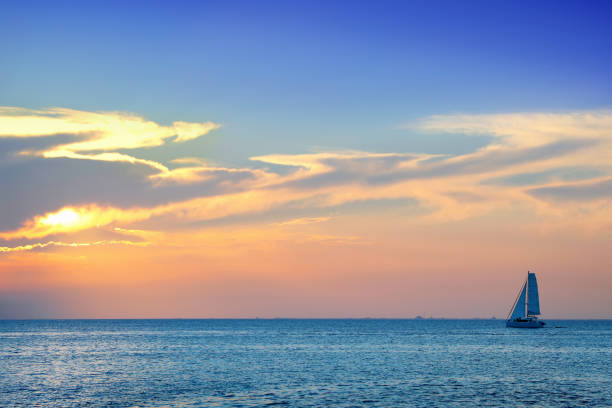 buntes meeresbild mit glänzendem meer und segelboot über trüben himmel und sonne bei sonnenuntergang - sailboat sunset nautical vessel sun stock-fotos und bilder