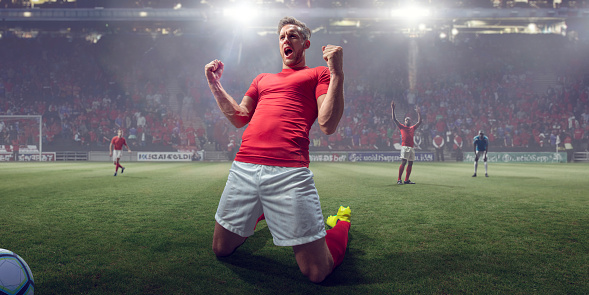 Close up image of a professional male soccer player kneeling on a pitch with fists clenched, arms raised, screaming with open mouth in celebration of victory. The footballer is wearing a plain red top and white shorts. Action occurs during a football game in a floodlit stadium full of spectators.