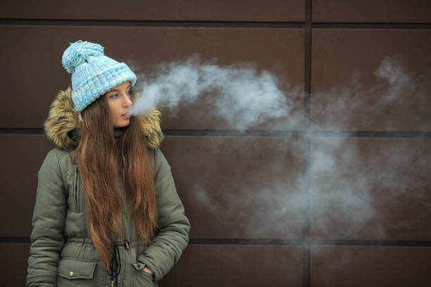 ・ vape ・ティーンエイジャー。冬の路上で近代的な茶色の背景の反対の電子タバコを吸っている若いかわいい白人の女の子。 - 電子タバコ ストックフォトと画像