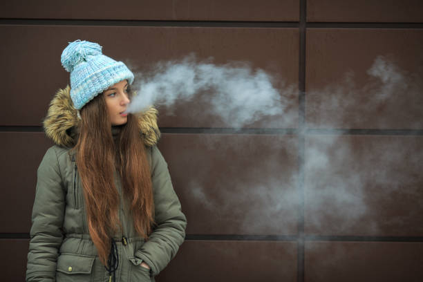 Vape teenager. Young pretty white girl smoking an electronic cigarette opposite modern brown background on the street in the winter. Vape teenager. Young pretty white girl in a cap smoking an electronic cigarette opposite modern brown background on the street in the winter. Bad habit. Close up. electronic cigarette stock pictures, royalty-free photos & images