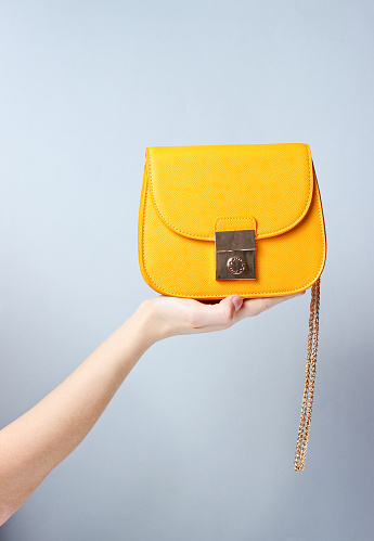 Female hand hold fashionable yellow leather bag with golden chain on gray background.