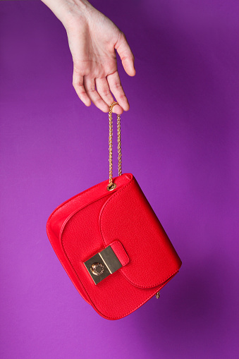 Female hand hold fashionable red leather bag with golden chain on purple background.