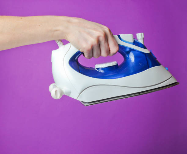 istock Female hand holding iron for ironing against purple background. 1153464368