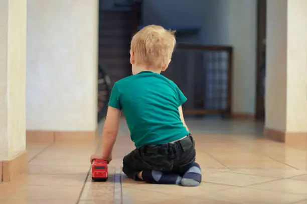 Photo of Little alone caucasian baby boy with fair hair sits on the floor, back to the viewer, with red bus toy. Loneliness concept.