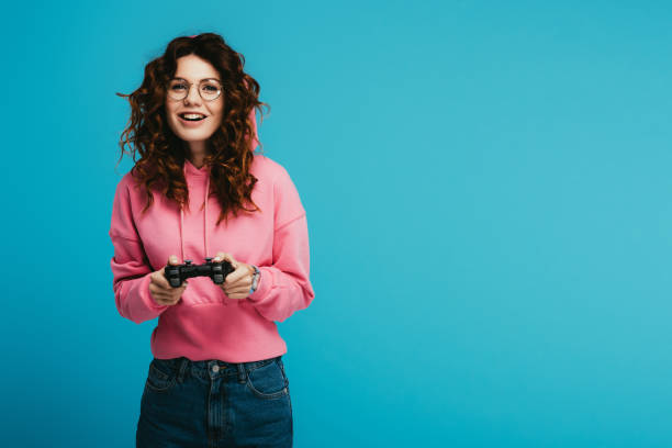 happy curly redhead girl playing video game while holding joystick on blue