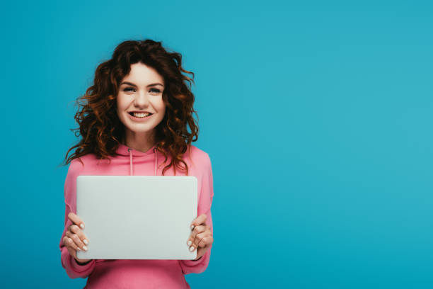 istock cheerful curly redhead girl holding laptop while standing on blue 1153470044