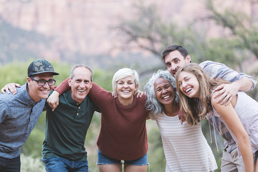 A multiethnic group of friends embrace and smile at the camera. They're lined up loosely and desert vegetation and sagebrush are in the background.