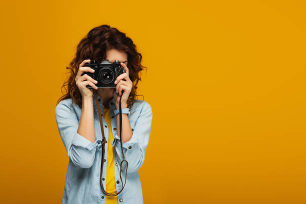 istock curly redhead photographer covering face with digital camera isolated on orange 1153476220