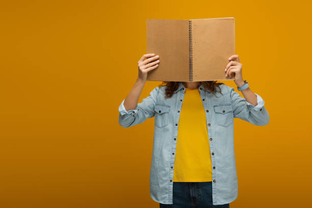 istock young woman standing and covering face with notebook on orange 1153476250