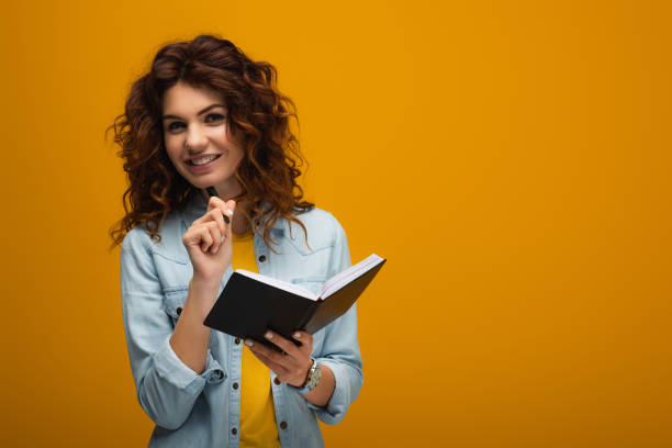 istock smiling redhead young woman holding notebook and pen on orange 1153476369