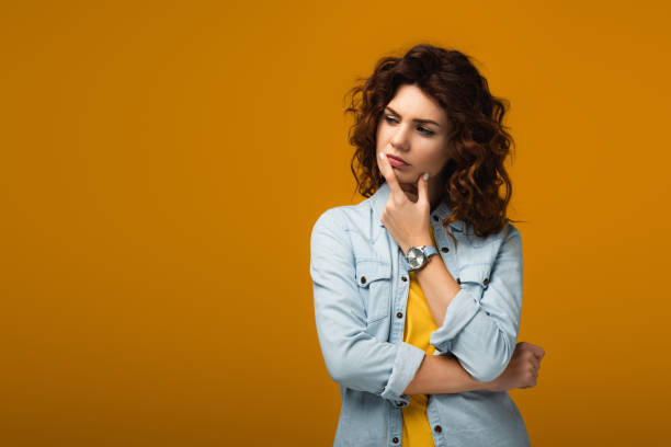 istock pensive curly redhead woman touching lips while thinking on orange 1153477680