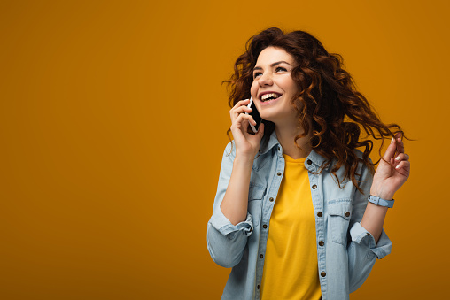 happy curly redhead woman talking on smartphone and touching hair on orange