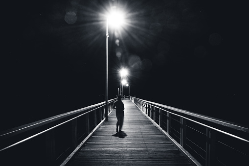 Silhouette of unrecognizable female walking along pedestrian bridge near street lamps in evening in Wellington Point, Australia
