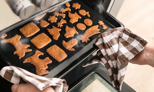 Golden brown baked Christmas cookies in decorative shapes on black tin and human hands with dish towels. Fresh Xmas pastries