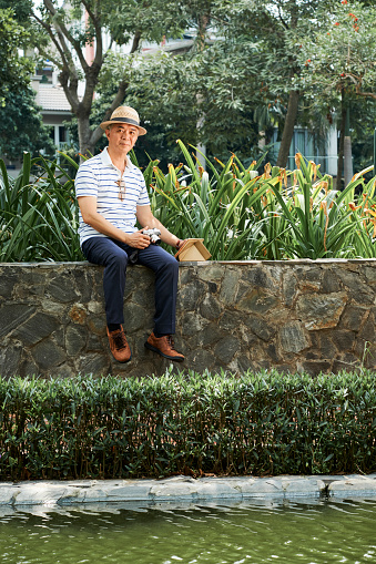 Portrait of Asian senior photograher sitting on stone wall near the pond and using photo camera and digital tablet after shooting the photos