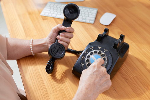 Close-up of mature woman sitting at office desk and dialing the number on retro phone