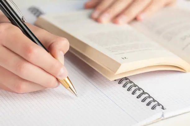 A man or teenager writes a black-yellow pen in a checkered notebook. The other hand is on the open book.