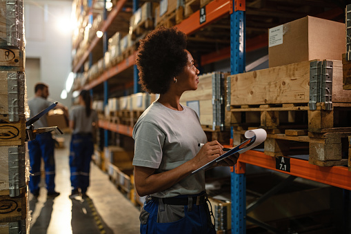 Black female warehouse worker going through delivery schedule while checking stock in storage room.