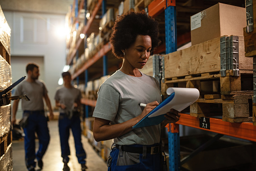 African American worker taking notes while making shipment schedule in a warehouse.