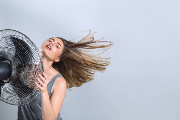 молодая женщина наслаждаясь прохладным ветром от электрического вентилятора - electric fan women wind beautiful стоковые фото и изображения