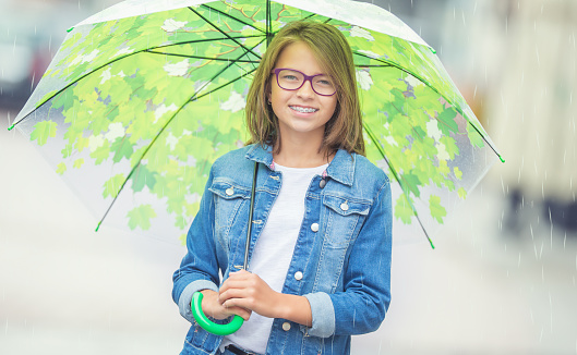 Portrait of beautiful young pre-teen girl with umbrella under spring or summer rain. Smilling girl with dental braces and glasses.