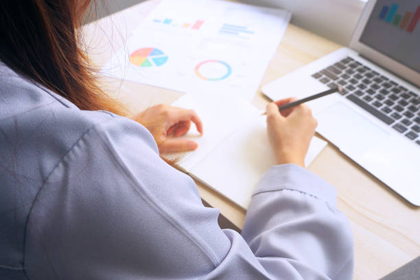 istock Working women write down on note book on working desk which have chart data paper and laptop,view from back. 1153586337