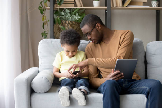 padre africano ayudando a pequeño hijo a explicar nueva aplicación o juego - african descent addiction african ethnicity rudeness fotografías e imágenes de stock