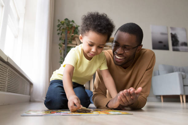 African father and little son collect jigsaw puzzle at home Black dad little son collect jigsaw puzzle put together pieces on warm floor in living room. Leisure activity have fun at home, logical reasoning ability, develop solving and fine motor skill concept piece of mind stock pictures, royalty-free photos & images