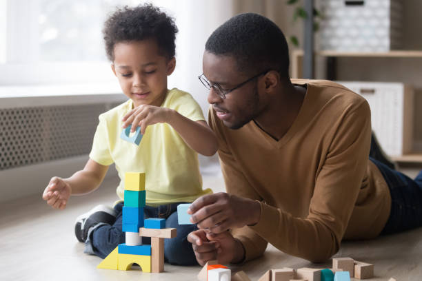 African father and son play with colourful toy blocks Little african boy building tower use colourful toy blocks set sitting on wooden warm heated floor in playroom with daddy or babysitter man, educational game, family at home leisure activities concept toy block photos stock pictures, royalty-free photos & images