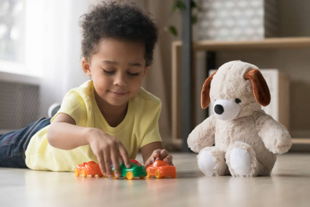 Adorable african kid lying on warm floor play with toys Mixed race adorable sweet toddler kid boy lying on warm heated wooden warm in modern home playroom playing with toy car and soft fluffy dog, children development and imagination creative games concept playroom stock pictures, royalty-free photos & images