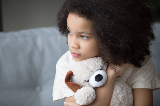 Unfortunate stray biracial kid girl embrace stuffed toy looking aside Close up upset stray biracial kid girl sitting on sofa cuddle stuffed toy dog looks aside feel lonely unhappy. Preschool age orphan, bullying discrimination, depression, psychological trauma concept pessimism photos stock pictures, royalty-free photos & images